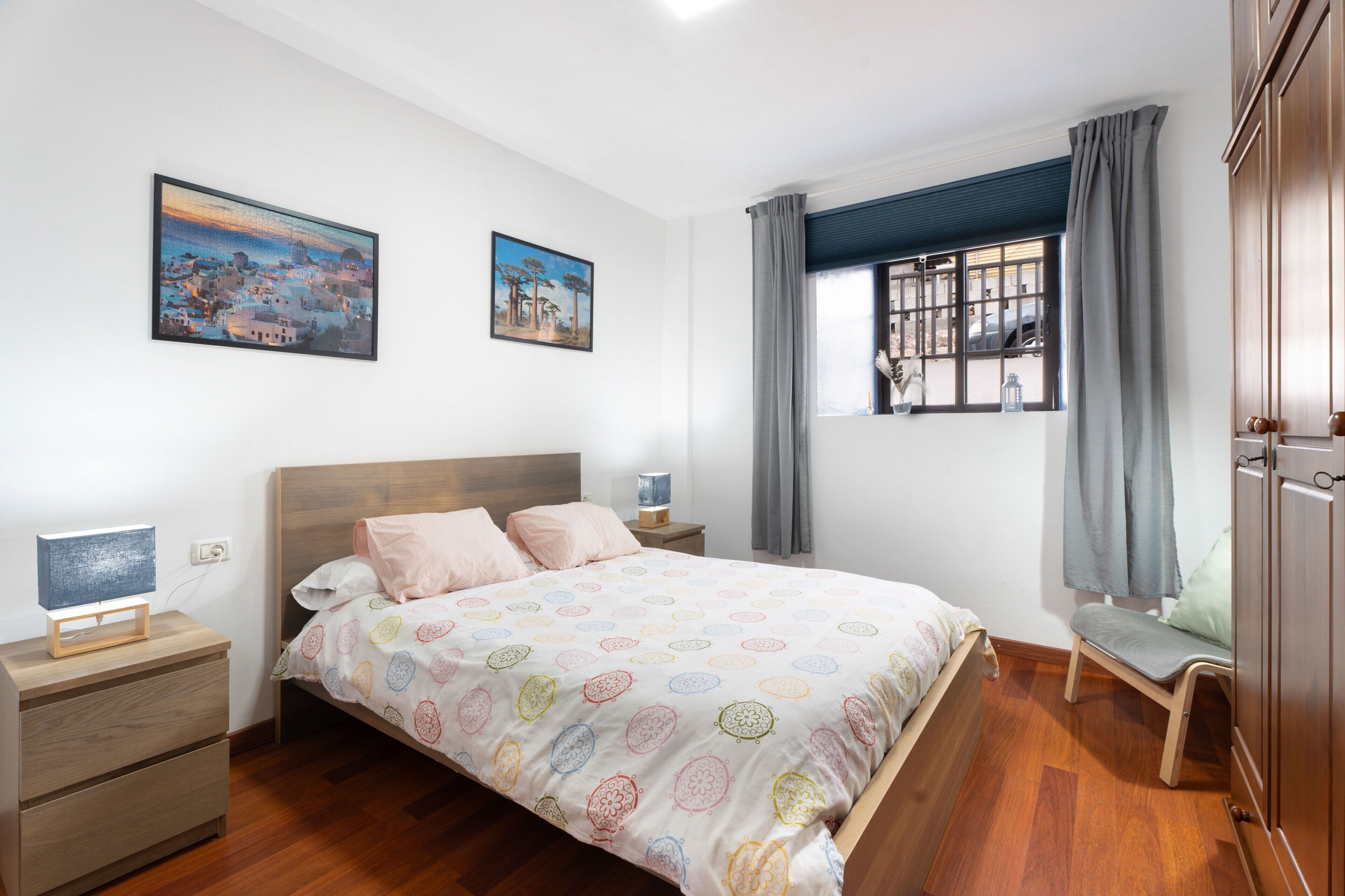 Un dormitorio con cama, mesitas de noche, una silla, un ventanal con cortinas y dos cuadros colgados en la pared.