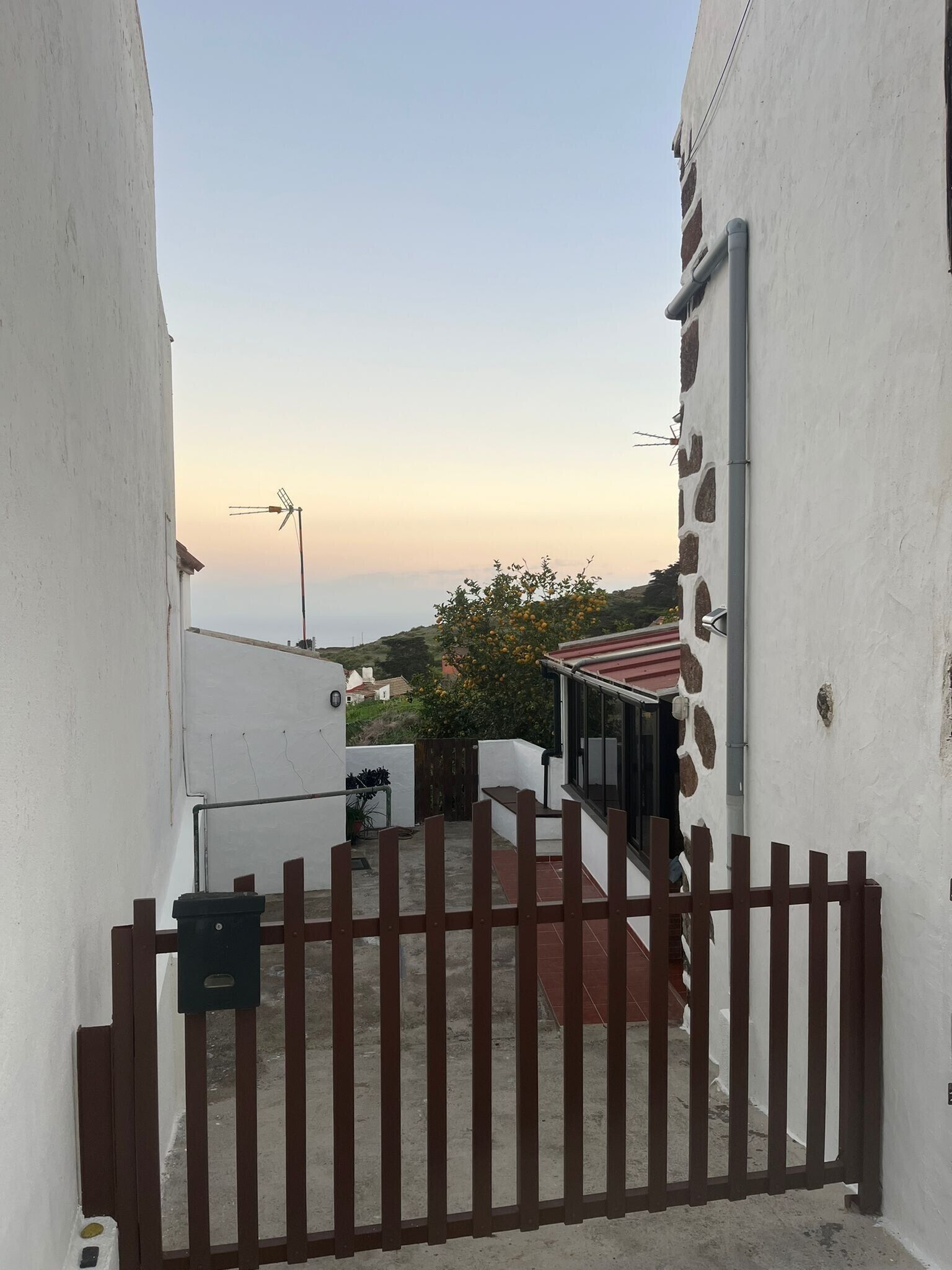 A narrow alleyway with a gated entrance, a small outdoor seating area, and a view of a distant landscape.