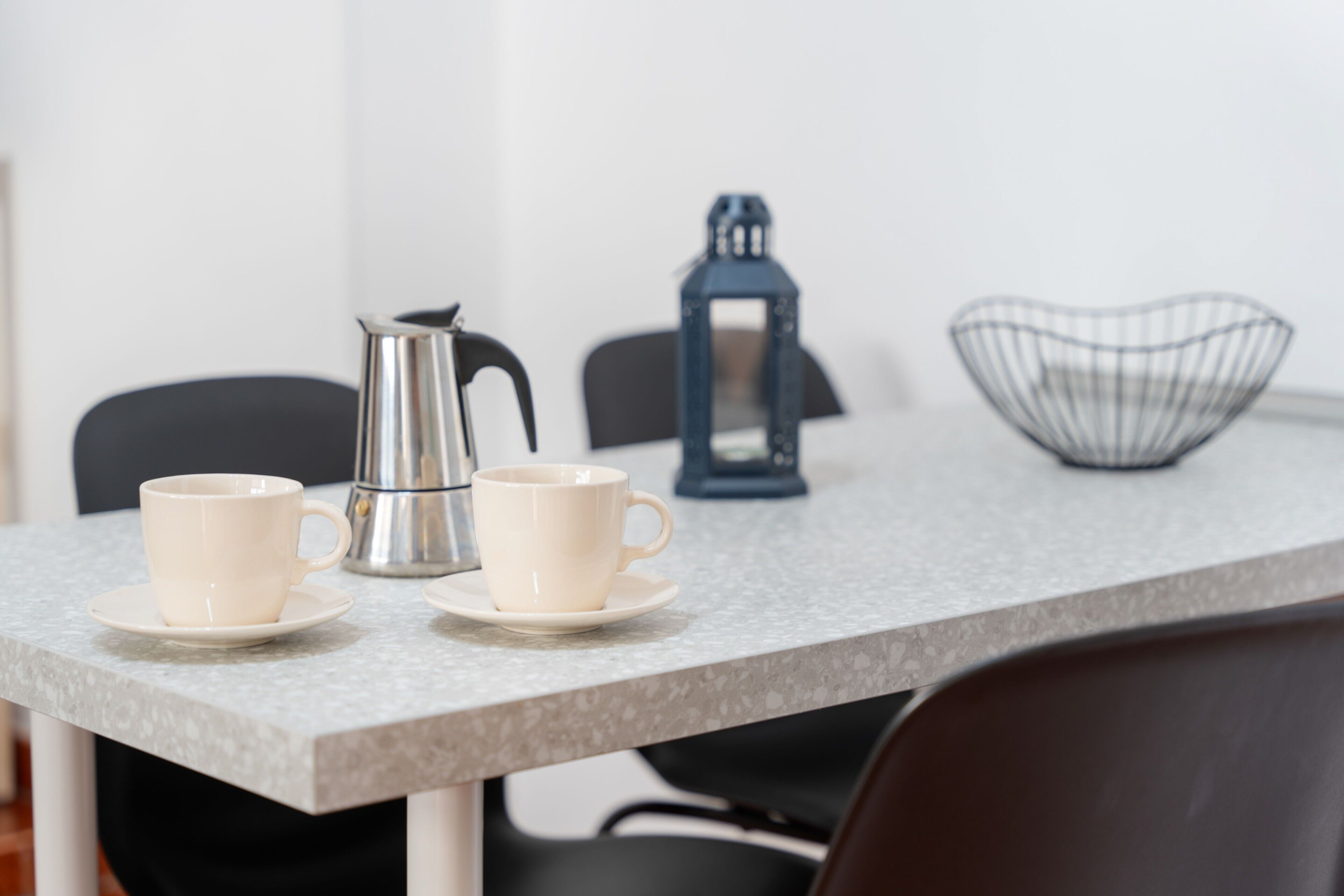 A table with two white cups and saucers, a silver coffee pot, a blue lantern, and a wire basket.