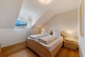 Quarto com teto inclinado, cama com cabeceira de madeira, mesa de cabeceira com abajur e janela com vista para montanhas.