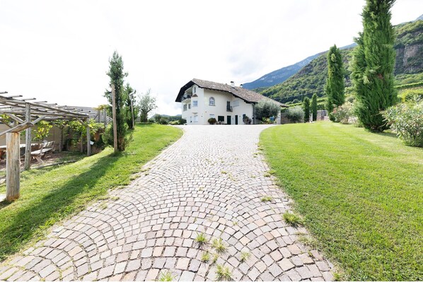 A paved pathway leading to a white building with a sloped roof, surrounded by greenery and mountains.