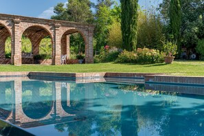 Una piscina con un arco de ladrillo, rodeada de follaje y un césped bien cuidado.