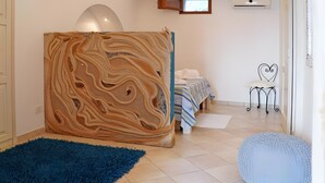 Ein Zimmer mit großformatiger abstrakter Wandkunst, einem Bett mit gestreifter Bettwäsche, einem blauen Teppich und einem kleinen runden Hocker.