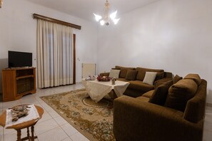 A living room with a sofa, a coffee table, a television on a stand, a chandelier, and a large window with curtains.