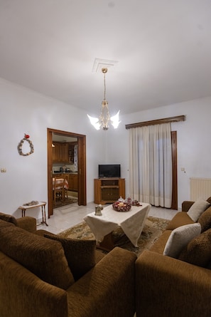 A living room with brown sofas, a table with a white tablecloth, a television, and a kitchen visible through an open door.