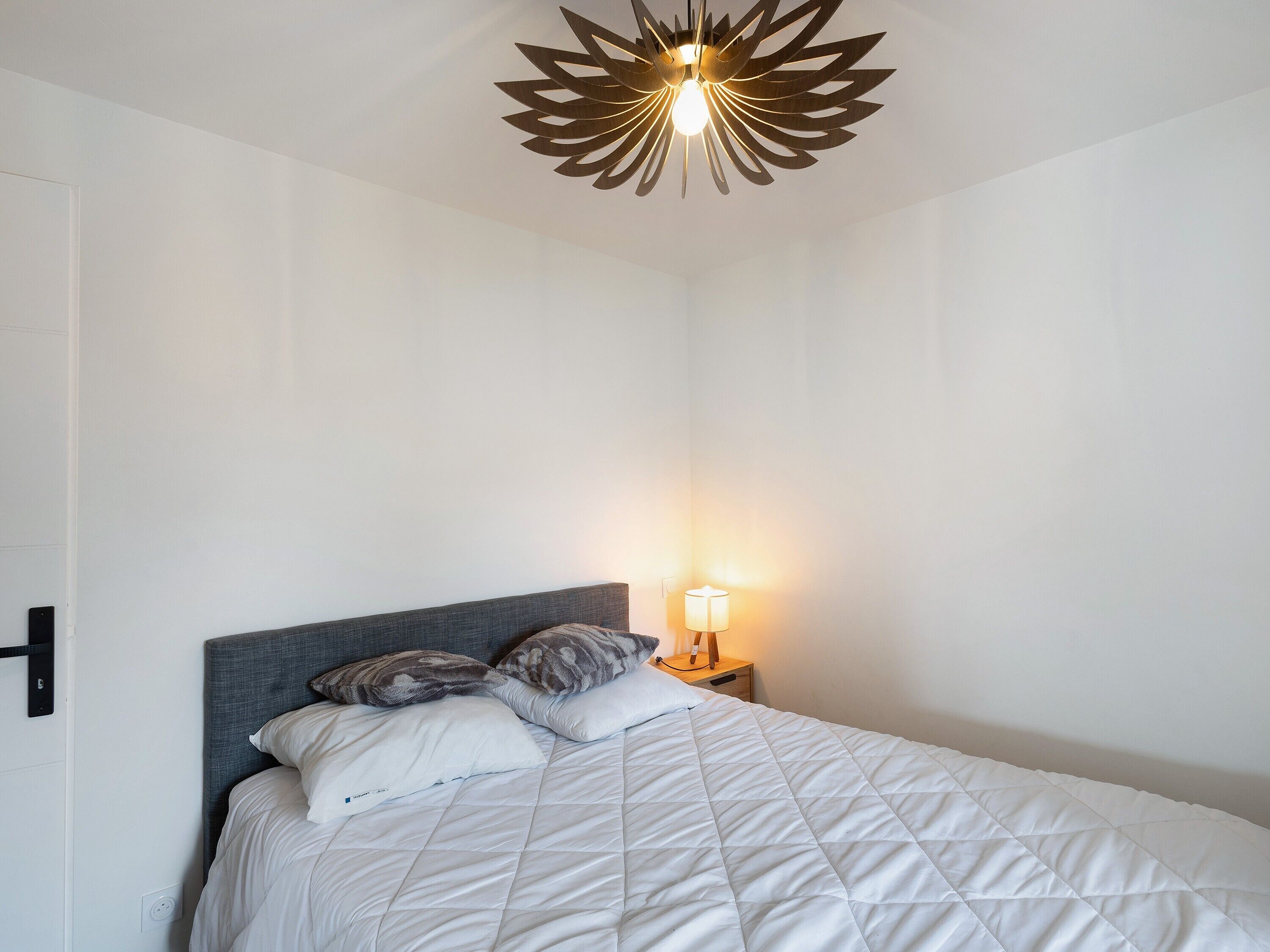 Ein ordentlich bezogenes Bett mit weißer Steppdecke, einem grauen Kopfteil und zwei Kissen. Auf einem hölzernen Nachttisch steht eine Nachttischlampe mit weißem Lampenschirm. Eine moderne, geometrische Deckenleuchte ist über dem Bett zu sehen.