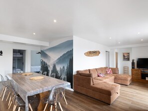 A modern living room with a wooden dining table, clear plastic chairs, a brown sofa, and a mounted TV. - Comfortable Mountain Stay in Formiguères: 4Bedrooms, 9Min from the Ski Resort with Private Terrace ! (Formiguères)