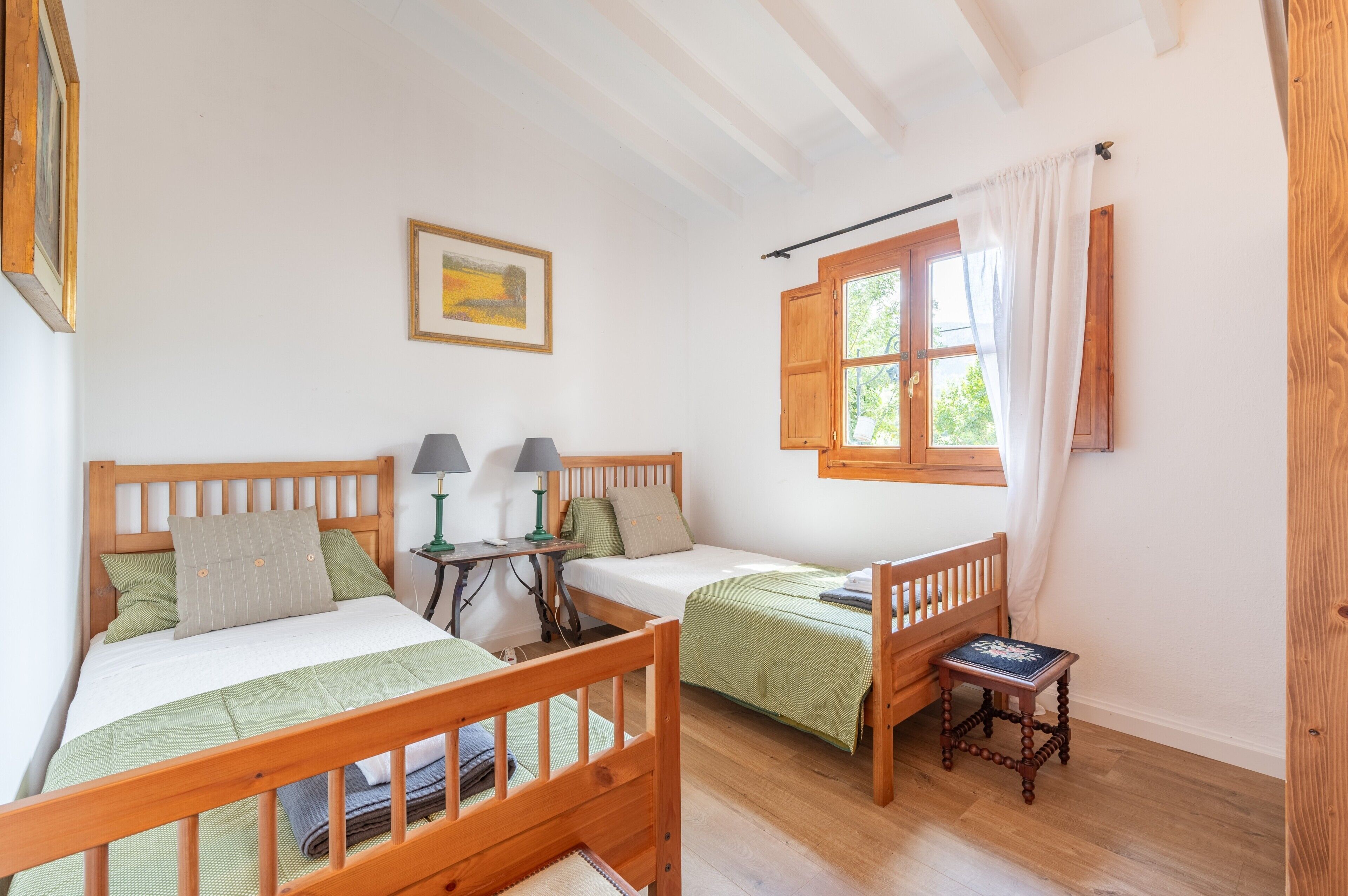 A room with two wooden beds, a small table with lamps, a window with curtains, and a framed picture on the wall.