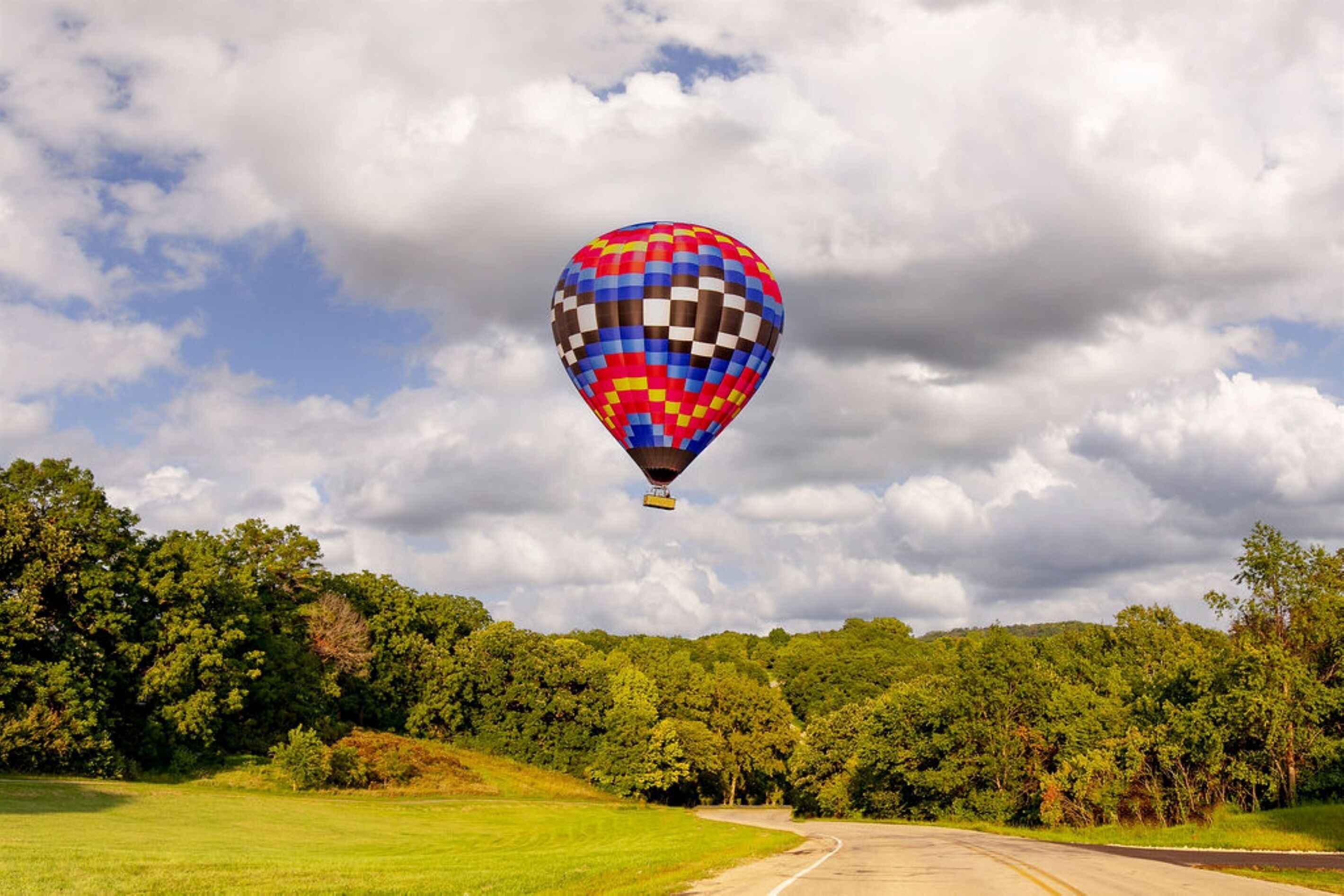 Une montgolfière à motifs en damier survole une route bordée d’arbres verdoyants.