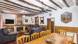 Un salon chaleureux avec une cheminée, des poutres apparentes, un téléviseur et un coin repas comprenant une table et des chaises en bois.