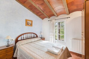 A bedroom with a wooden headboard, a bed with a patterned bedspread, a nightstand with a lamp, a window with curtains, and a radiator.
