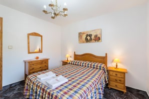 A bedroom with a bed, a wooden nightstand, a bedside table, a mirror, a chandelier, and a painting on the wall. - Apartment 'Villacalar Il' with Mountain View, Private Garden and Wi-Fi (Calar de la Santa)