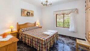 A bedroom with a bed, a nightstand, a lamp, a window with curtains, and a painting on the wall. - Apartment 'Villacalar Il' with Mountain View, Private Garden and Wi-Fi (Calar de la Santa)