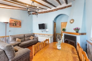 Uma sala de estar com uma mesa de jantar de madeira, sofás, lareira e uma televisão fixada na parede.