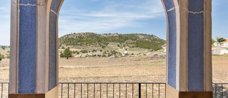 A view through an arched window with a metal railing, overlooking a hilly landscape with sparse vegetation and buildings in the distance.