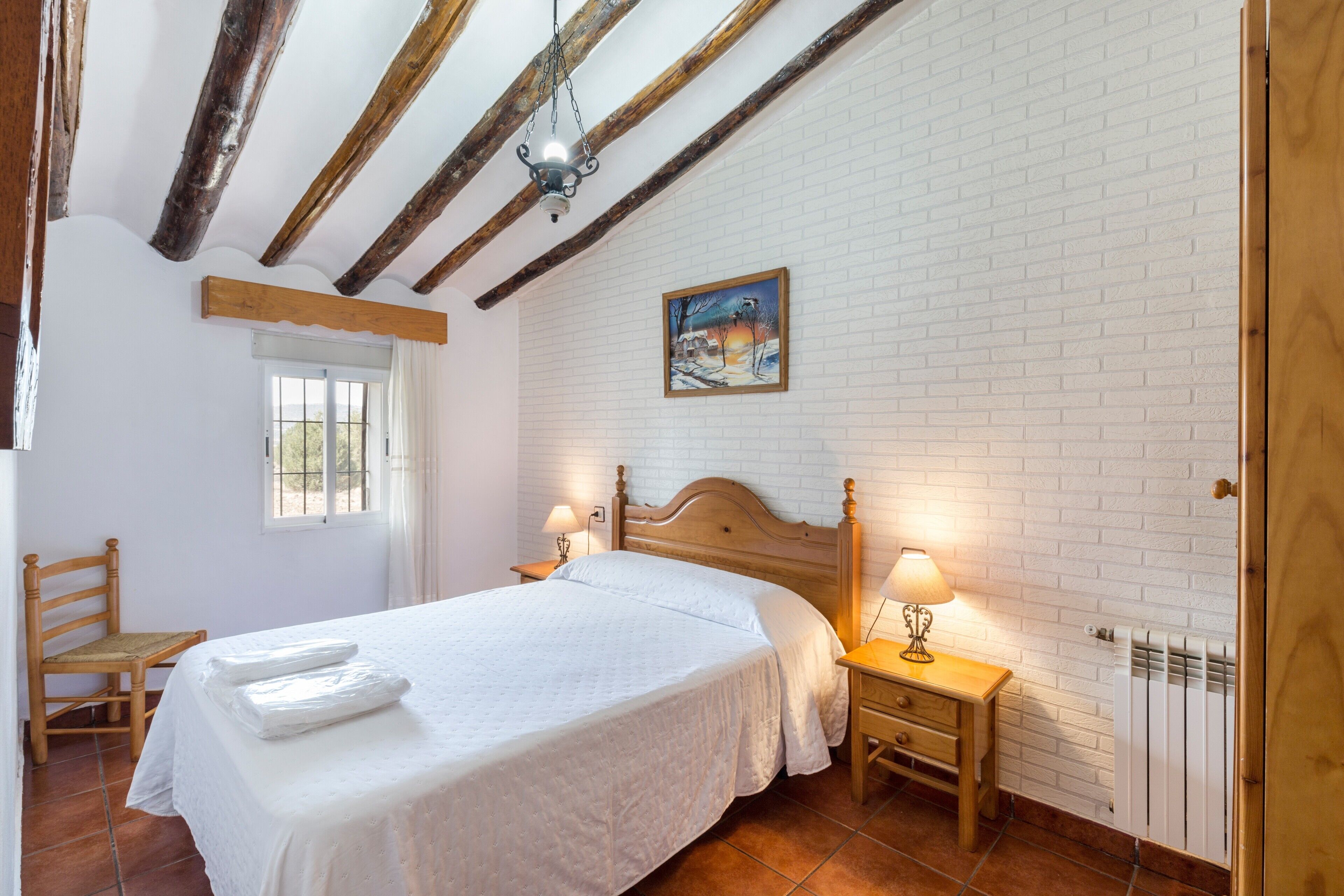 Une chambre à coucher avec un lit, une tête de lit en bois, deux tables de chevet avec des lampes, une fenêtre avec des rideaux, une chaise en bois et un mur en briques blanches.