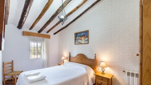 Un dormitorio con cama, cabecera de madera, dos mesitas de noche con lámparas, ventana con cortinas, una silla de madera y una pared de ladrillo blanco.