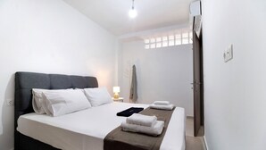Cama bem arrumada com lençóis brancos e manta marrom, luminária de cabeceira e janela com cortinas.
