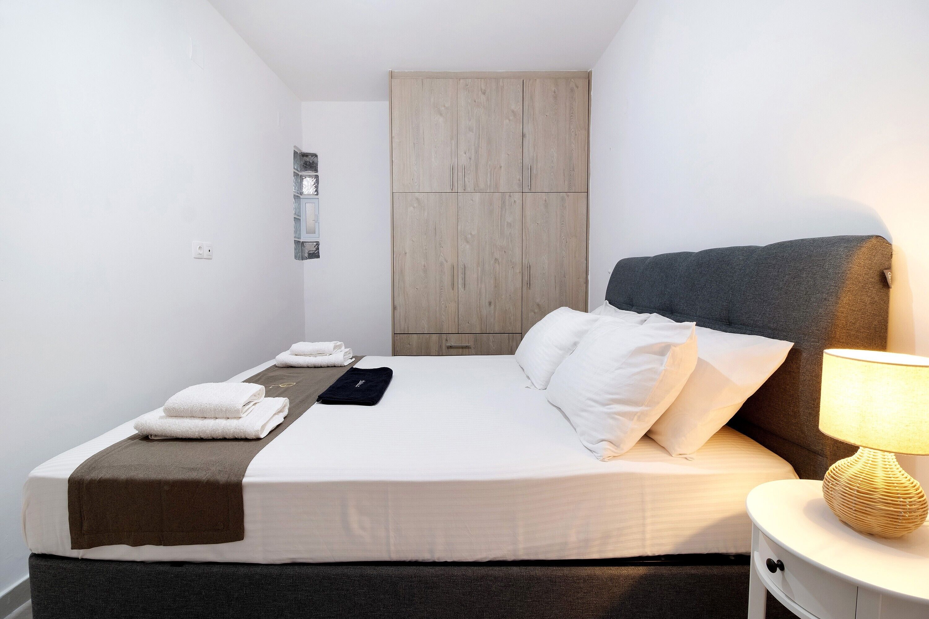 Una cama bien tendida con almohadas, una lámpara de noche y una toalla doblada sobre una mesita blanca.