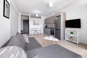 Una sala moderna con un sofá gris, una mesa de comedor con sillas, una zona de cocina con refrigerador y un televisor en la pared.