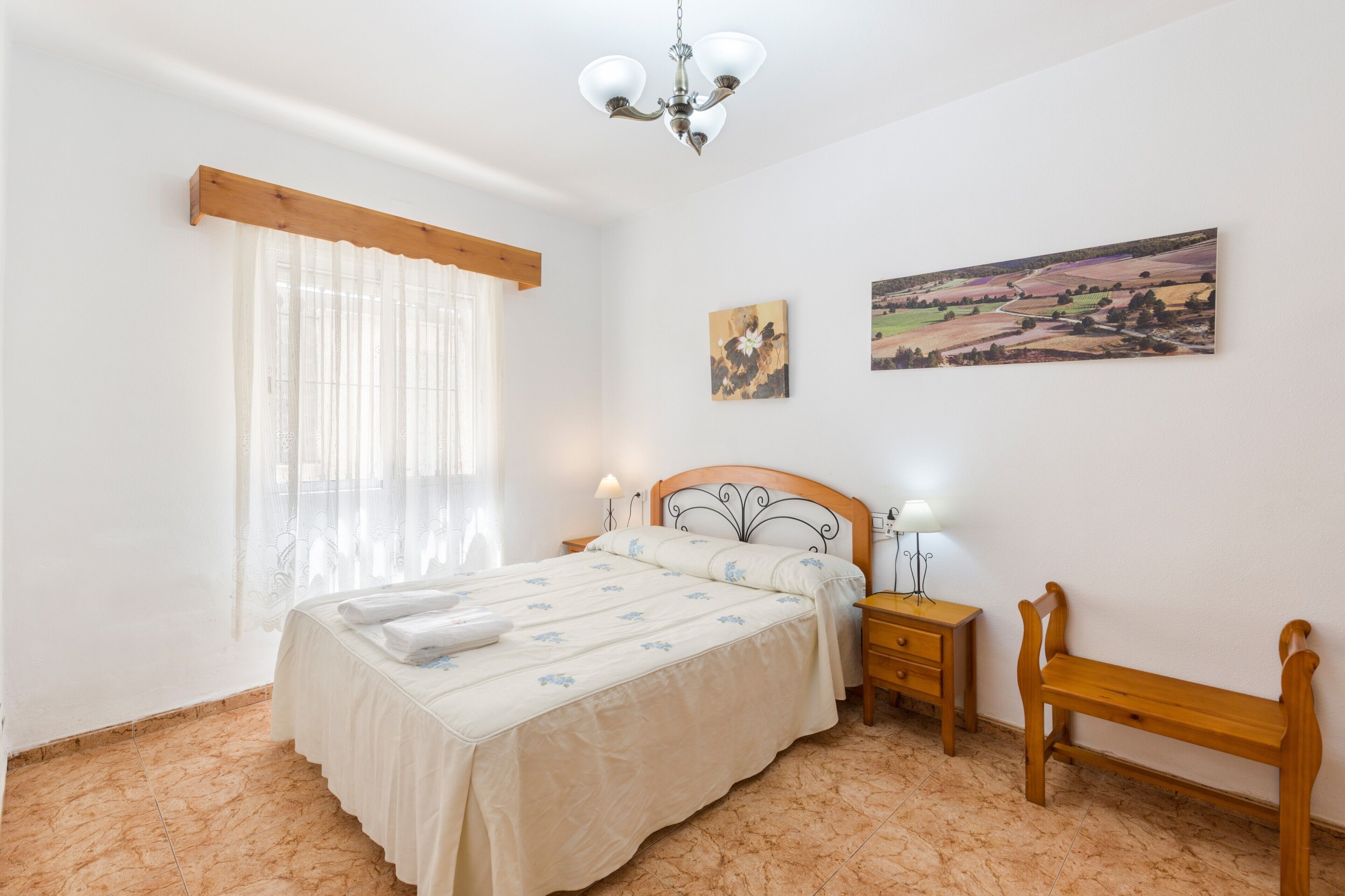 A bedroom with a bed, bedside tables, a bench, a chandelier, and two landscape paintings on the wall.