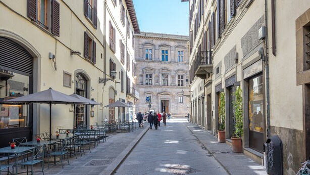 Una calle estrecha con mesas al aire libre, un edificio con puerta azul y cielo despejado.