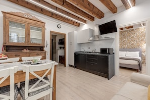 Una cocina moderna con vigas de madera, un comedor con mesa y sillas blancas, y un dormitorio con cama y papel tapiz estampado.