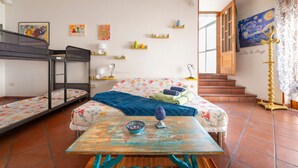 A room with a bunk bed, a single bed with a floral bedspread, a table with a bowl and a decorative item, a staircase leading to an upper floor, and a painting on the wall.