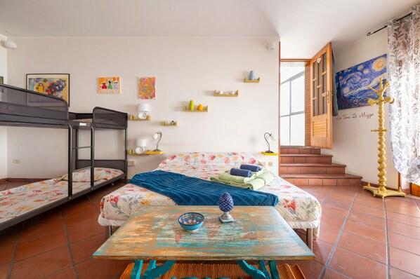A room with a bunk bed, a single bed with a floral bedspread, a table with a bowl and a decorative item, a staircase leading to an upper floor, and a painting on the wall.