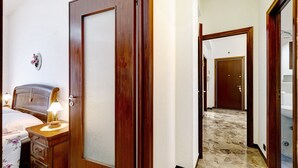 Ein Hotelzimmer mit Holztür, einem Nachttisch mit Lampe, einem Bett mit Blumenmuster-Bettdecke und einem durch eine offene Tür einsehbaren Badezimmer.