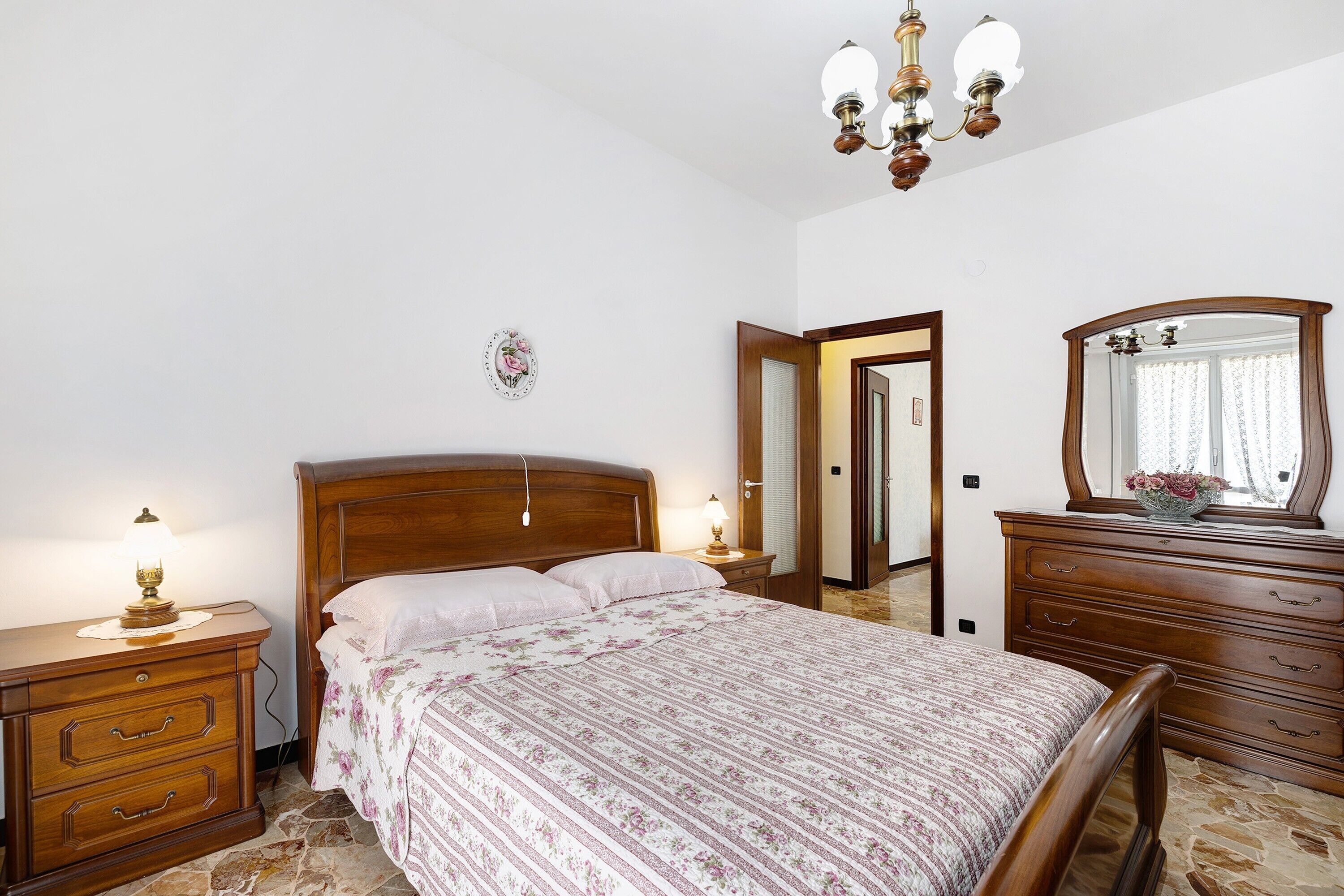 A bedroom with a wooden bed, bedside tables with lamps, a dresser, a mirror, and a door leading to another room.