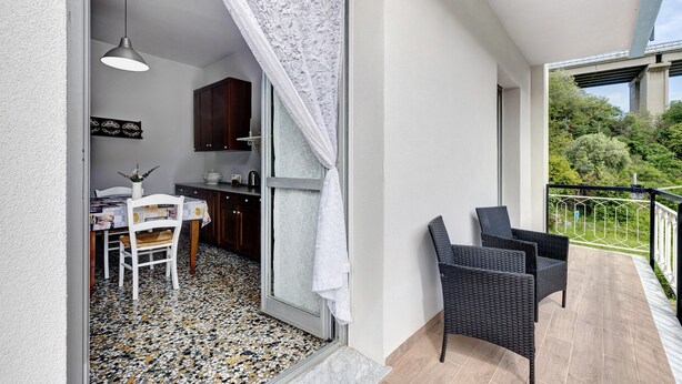 Ein Balkon mit Korbstühlen, eine Essecke mit Tisch und Stühlen sowie eine durchgehende Tür sichtbare Küche.