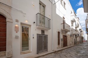 Uma rua estreita de calçada, flanqueada por edifícios brancos, cada um com uma varanda e uma porta com persianas.