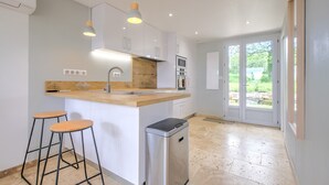 A modern kitchen with a central island, bar stools, a built-in oven, and a trash bin. - Gîte Déodatus in Cladech – House with Garden, Saltwater Pool, and Air Conditioning (Cladech)