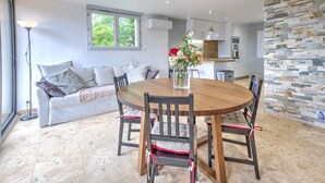 A modern dining area with a round wooden table, cushioned chairs, a sofa, and a stone wall. - Gîte Déodatus in Cladech – House with Garden, Saltwater Pool, and Air Conditioning (Cladech)