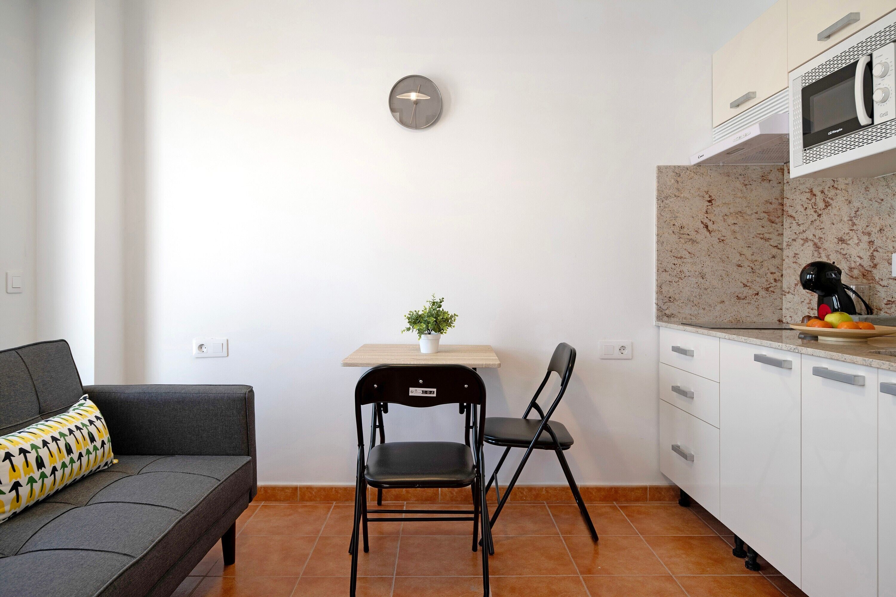 A compact kitchen with a microwave, a small table with a plant, a sofa, and two chairs.