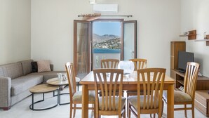 A modern living room with a sofa, dining table, chairs, a TV, and a balcony with a view of a mountainous landscape. - Apartment 'Akrogiali House Drimonas' with Sea View, Wi-Fi and Air Conditioning (Leros)