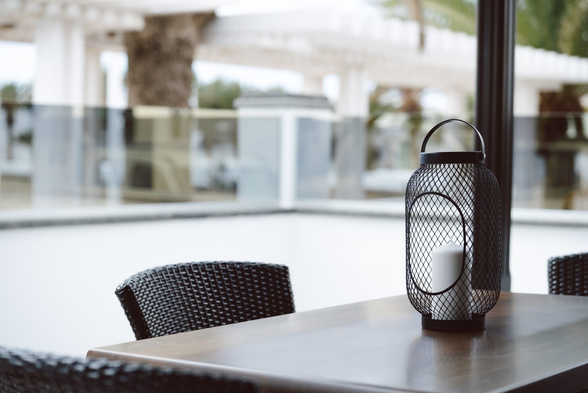 Una linterna de metal sobre una mesa con sillas de mimbre, una barandilla de vidrio y vistas a un edificio y árboles.