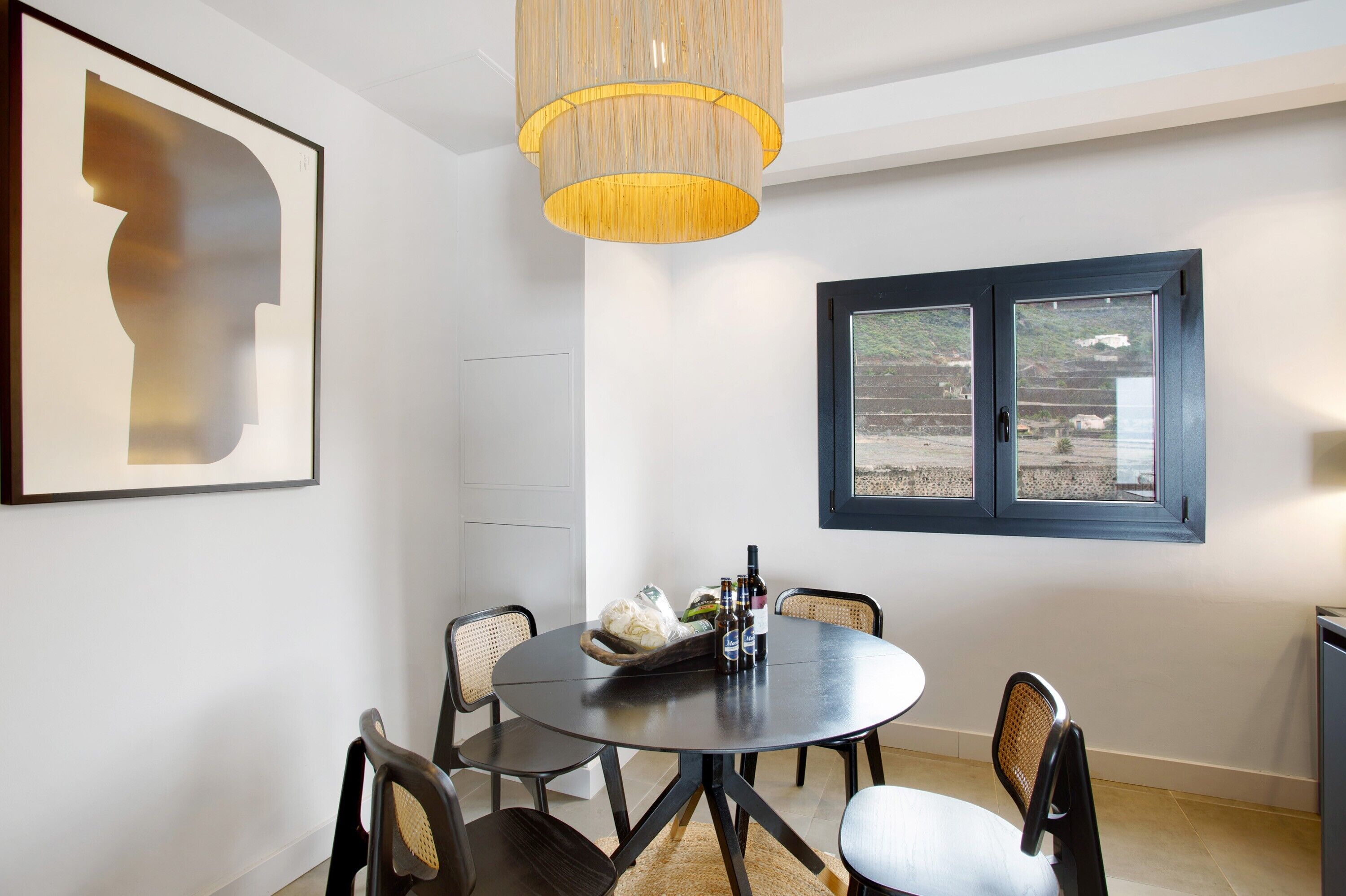 Un comedor con una mesa redonda, sillas, una lámpara colgante, una obra de arte enmarcada y un ventanal con vistas.