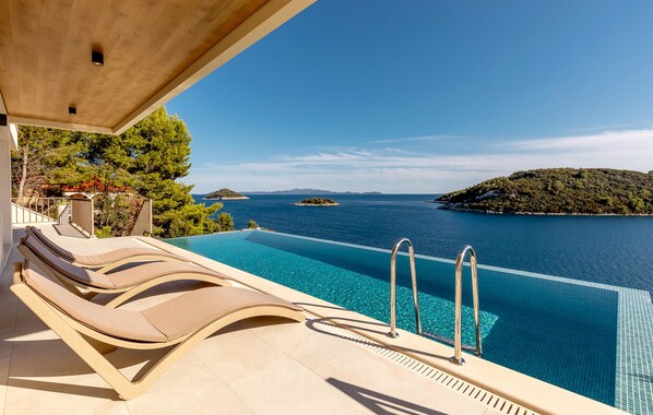 Une piscine à débordement avec des chaises longues, offrant une vue imprenable sur une mer d’un bleu cristallin et sur des îles.