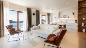 Una sala de estar moderna con un sofá blanco, dos sillones marrones, una mesa de centro de vidrio y vistas al mar a través de grandes ventanales.