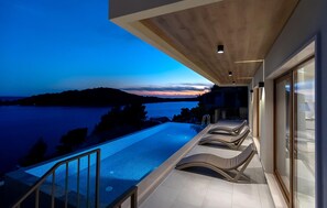 Une piscine à débordement moderne offrant une vue sur un coucher de soleil se reflétant sur l’eau, un plafond en bois et une porte vitrée donnant sur un balcon.