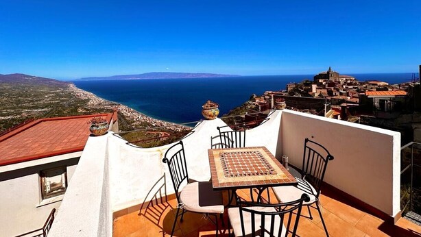 A terrace with a table and chairs overlooking a coastal town and the sea. - Holiday Home 'Casa Mark' with Sea View, Wi-Fi and Air Conditioning (Forza d'Agrò)