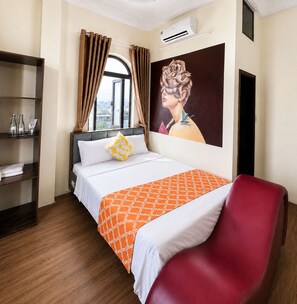 A hotel room with a bed, a red chair, a window with curtains, an air conditioning unit, and a painting on the wall.