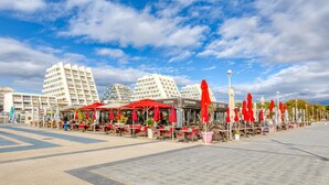 Una plaza pública con mesas al aire libre, sombrillas rojas y edificios al fondo.