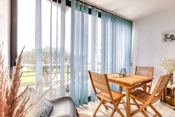 A dining area with a wooden table and chairs, a view through large windows with blue curtains, and a white textured wall.