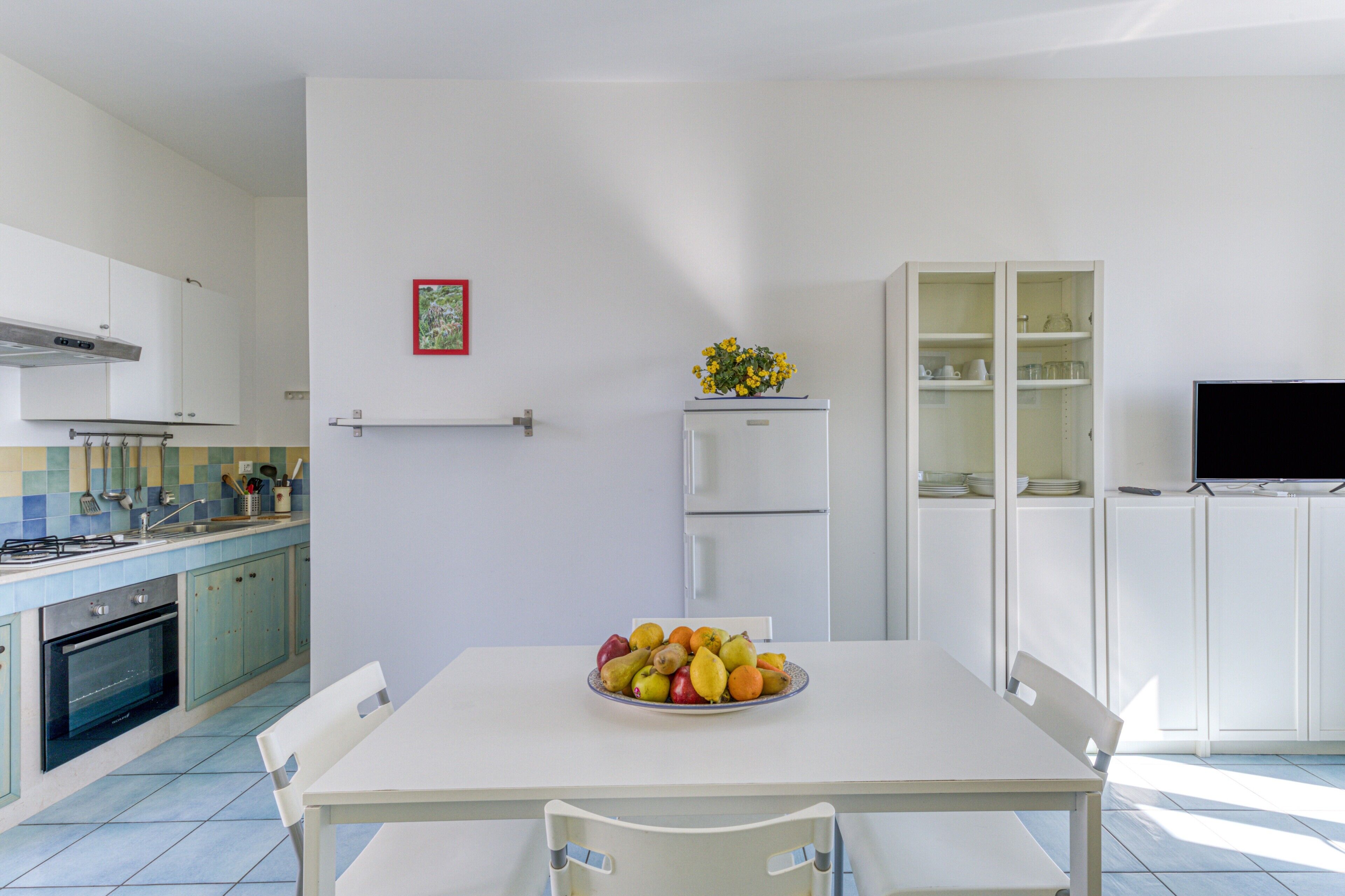 Eine moderne Küche mit Esstisch, Stühlen, einer Obstschale, einem Kühlschrank und einem Fernseher.