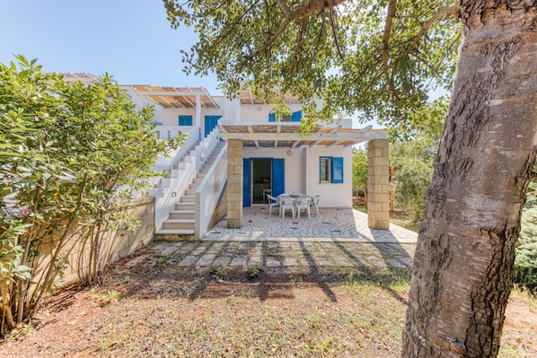 A two-story building with a white exterior and blue shutters, a stone staircase, and a patio area with a table and chairs.