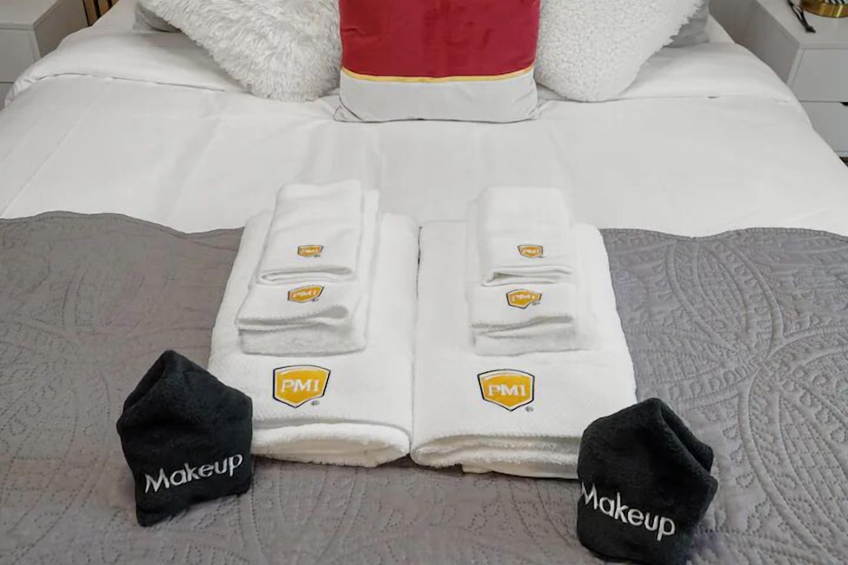 A neatly made bed with white linens and two towels folded on top, each with a 'PMI' logo. Two black 'Makeup' bags are placed on the bed.