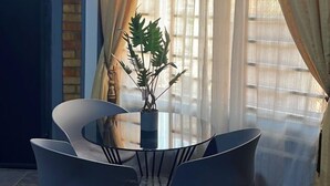 A dining area with a round table and chairs, a potted plant, and a window with sheer curtains. - Cozy Bricks Apartment (Obomeng)
